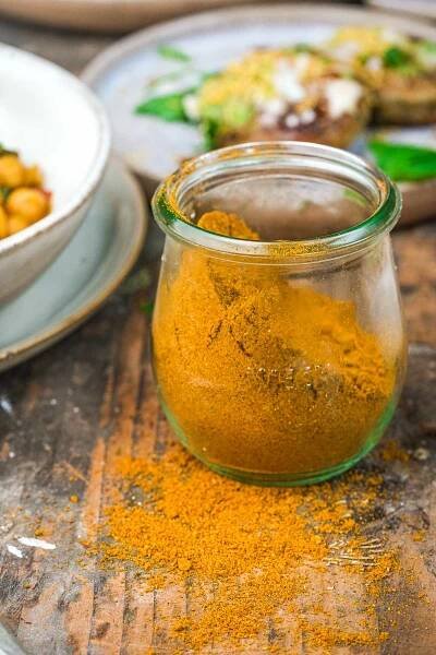 A small glass jar filled with Madras Curry Powder sits on a wooden surface, with some spilled around the jar. Plates with food are in the background.