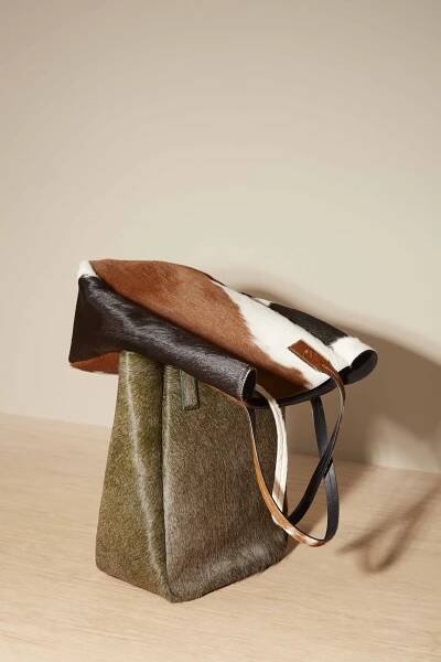 Two calf-hair tote bags - one olive green and other burnt orange and white sits on top. They are on a light wooden surface.
