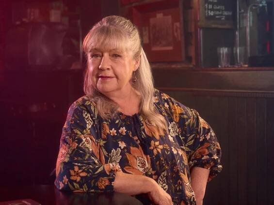 noni Hazlehurst, with fair long hair and fringe, sits near a front bar at a pub, smiling with closed mouth.