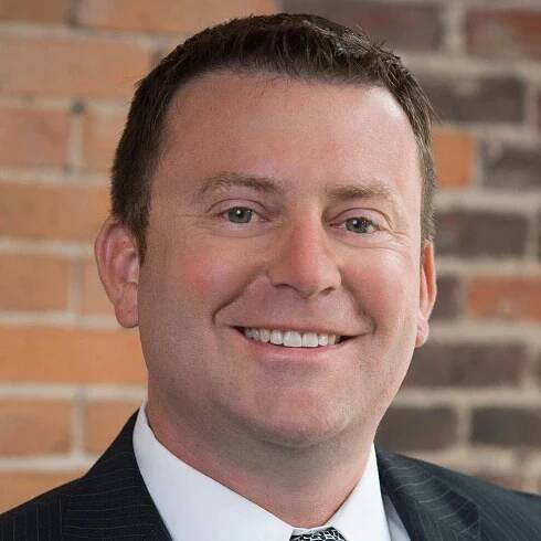 A man in a suit and tie is smiling in front of a brick wall.