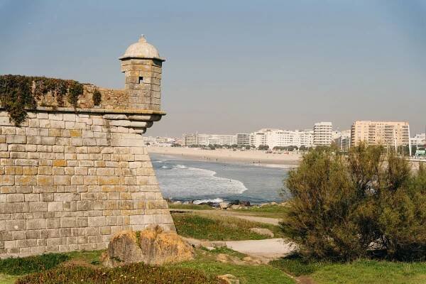 On the left in the foreground is an old pale stone fort with little lookout tower. In the background is a white sandy beach and ocean, with apartment blocks overlooking the beach.