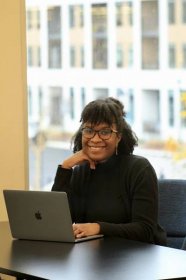 Black woman with curly black hair and wearing a black turtleneck and sweater sitting in front of an open laptop