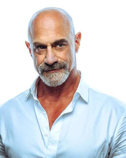 Chris Meloni poses against a plain backdrop with a bald head and graying beard wearing a white shirt