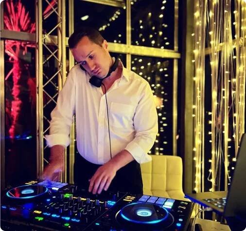 DeinDJ arbeitet an einem beleuchteten DJ-Pult bei Nacht, mit glitzernden Lichterketten im Hintergrund.