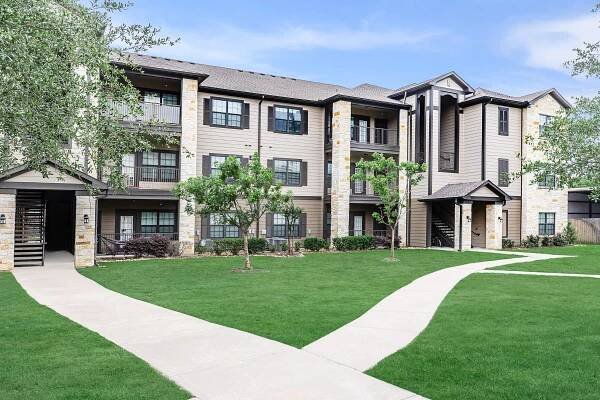 Modern apartment building with a blend of stone and wood siding, featuring multiple balconies and large windows. The well-maintained green lawns and decorative trees surround the building, and a paved pathway leads to the entrance. A clear blue sky provides a bright backdrop.