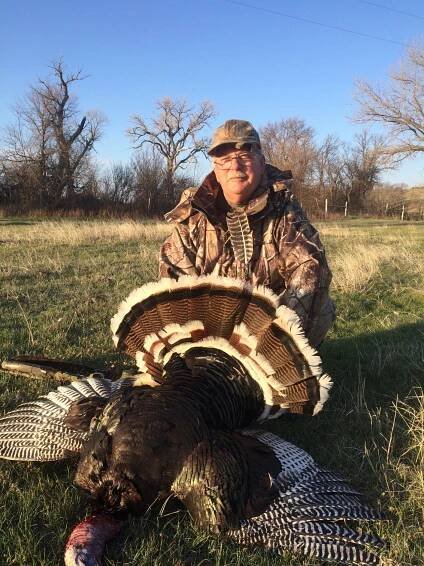 Man kneels next to a harvested turkey in a field. He wears camouflage. Turkey's feathers fanned out.