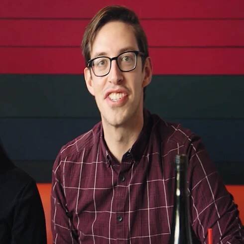 Person wearing glasses and a plaid shirt is seated indoors, looking towards the camera with a bottle in the foreground