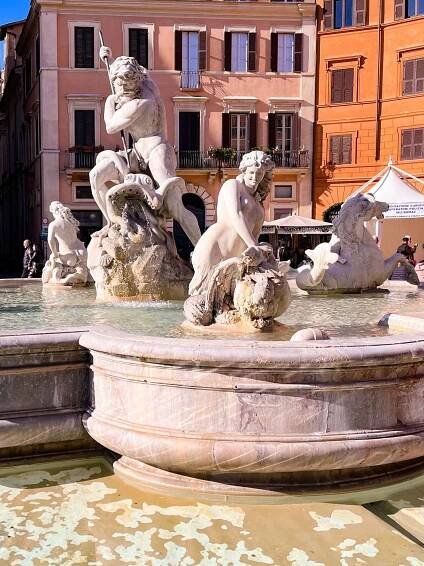 The photo shows the Fontana del Nettuno in Rome's Piazza Navona which features Neptune battling a sea creature, surrounded by nymphs. Marble statues shine under the sun, with cascading water accentuating their dynamic forms. The historic square's pastel buildings provide a charming backdrop to this emblematic work of Baroque art, capturing the essence of Roman heritage in the bustling heart of the city.