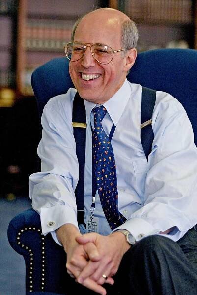 A middle-aged, smiling Michael Cardozo, wearing a blue shirt, dark pants, suspenders and a patterned tie, sits in an armchair, his hands clasped.