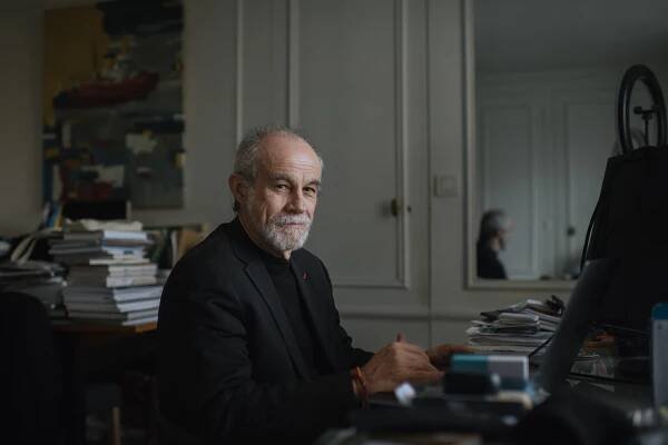Carlos Moreno, wearing a dark suit, sits in a room among piles of books and papers.