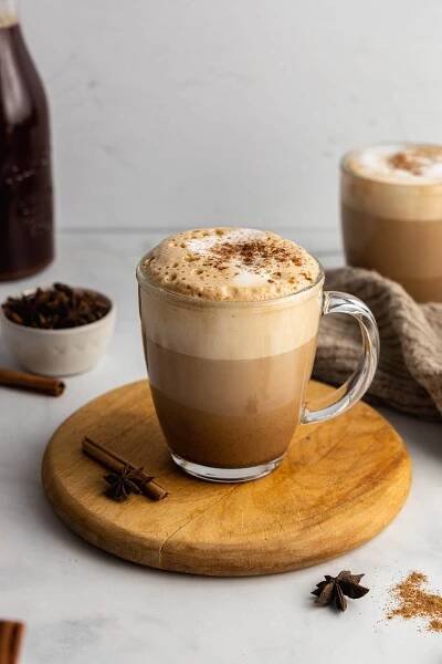 A clear glass mug filled with dirty chai latte garnished with chai spice sits on a round, wooden board.