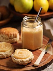 Portrait shot of Easy Quince Curd in a jar (with teaspoon) on a board with buttered scones topped with curd, knife and quinces in a wooden bowl in background and more scones..