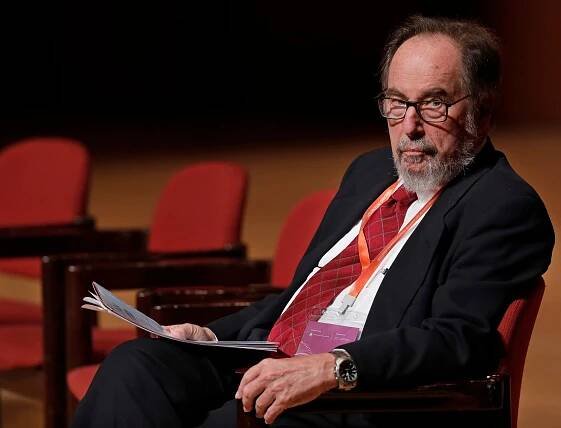 David Baltimore, a bearded man in glasses and a dark suit, sits in a red chair in what appears to be an auditorium.