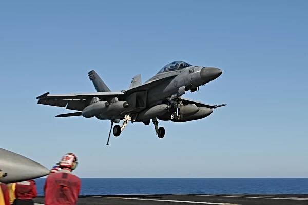 A fighter jet flying just above the deck of an aircraft carrier on the water. 
