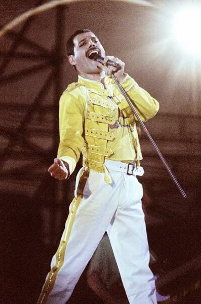 A singer passionately performs on stage, wearing a military-style jacket and white pants, holding a microphone with a spotlight behind them