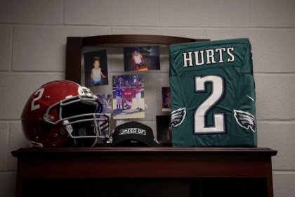 A red Alabama football helmet with the No. 2 on it; a black hat with a logo reading “Breed of 1”; a green Jalen Hurts Eagles jersey with the No. 2 on it; and family photos rest on top of a bookcase against a white wall.
