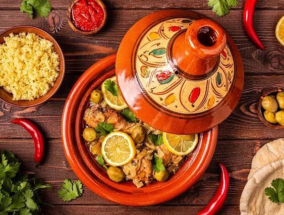 Morocco: A Moroccan tagine dish with chicken, olives, and lemon slices in a decorated clay pot, surrounded by couscous, chili peppers, cilantro, bread, and a bowl of red sauce on a wooden table.