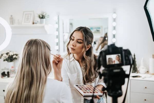 A makeup artist applies makeup to a client while filming a beauty vlog.