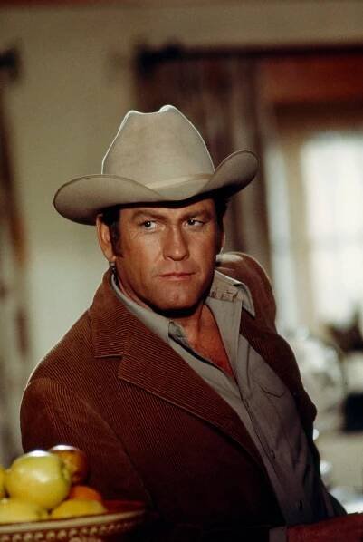 Earl Holliman, wearing a beige cowboy hat, brown corduroy jacket and gray shirt, leans up against a table or bar, next to a bowl of fruit.