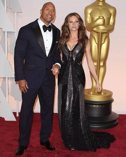 Dwayne Johnson looks dapper in a classic navy tuxedo, while Lauren Hashian stuns in a shimmering black gown with metallic accents, making a glamorous appearance at the Academy Awards