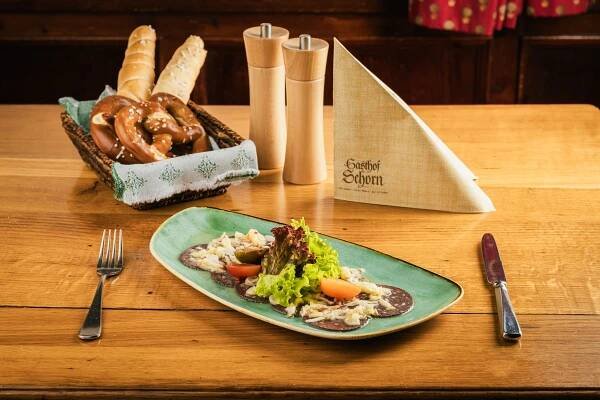 Holztisch mit grüner Platte voller Aufschnitt, Salat und Gemüse, daneben eine Besteckgarnitur, Brot und Brezeln im Korb.