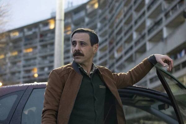 Kenny (Emun Elliott) leans on an open car door in front of a large housing block