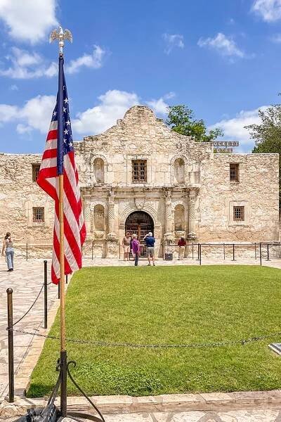 The green field in front of The Alamo with people walking and the American flag in the foreground, a significant stop on your Texas Hill Country road trip.