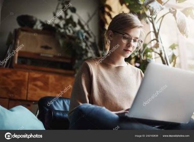 Horizontalshot of creative and stylish urban female student in glasses, sitting on crossed feet and holding laptop, looking at keyboard while typing, preparing homework or writing essay in cozy area Stock Photo by ©ringsaroundme 242162638