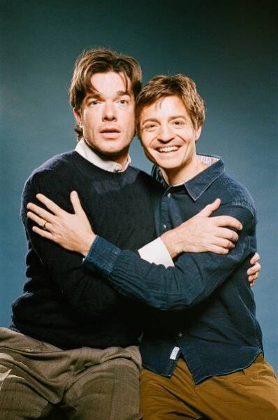 John Mulaney, in a navy blue sweater, and Simon Rich, in a button-up denim jean shirt, are sitting side by side and looking forward while embracing. 