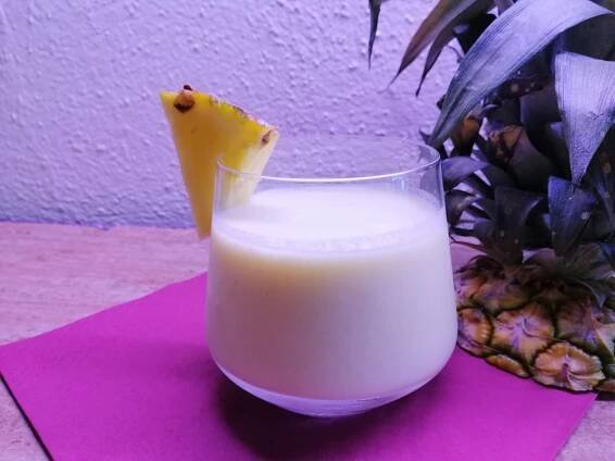 Ein Glas mit cremigem, gelbem Drink, garniert mit einem Ananasstück auf einem lila Serviette, neben dem Anschnitt einer Ananas. 