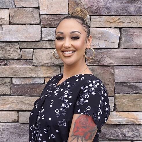 A woman with a tattoo on her arm is smiling in front of a stone wall.