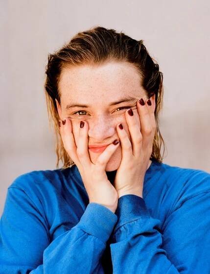 Frau mit blauer Bluse hält ihr Gesicht lächelnd mit beiden Händen.