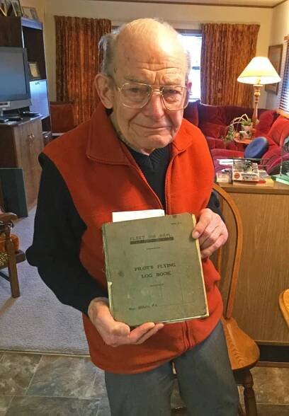 Elderly balding man with glasses and an impish smile, wearing a red fleece vest, holds a rather tattered green journal with the title on the front reading PILOT'S FLYING LOG BOOK.