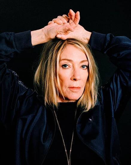 A woman with chin-length blond hair, dressed in black, brings both hands up above her head.