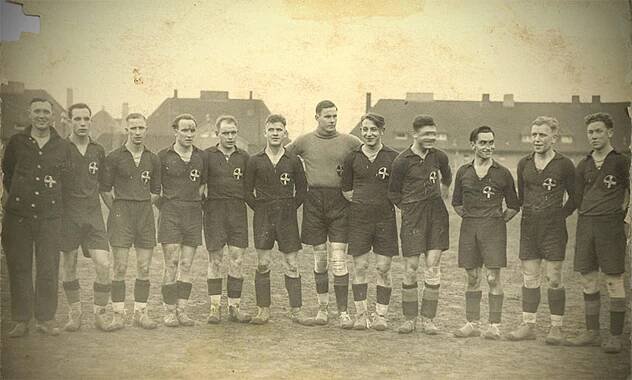 A vintage black-and-white photo of an 11-man football team in matching dark jerseys and shorts, standing in a line with their arms around each other’s shoulders. They are outdoors on a dirt field with buildings in the background.