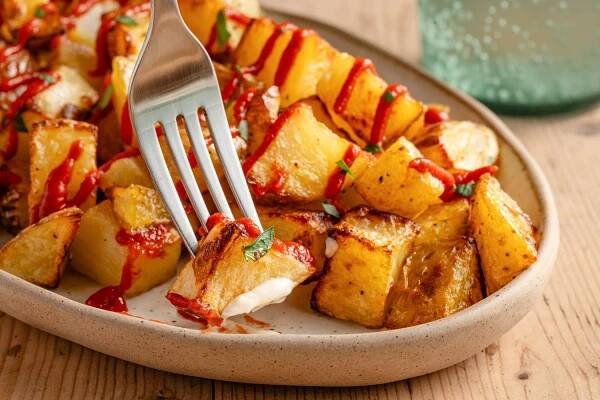 Closeup view of a piece of patatas bravas on a fork along with a platter of roasted diced potatoes topped with tomato sauce and parsley all on a wooden table top