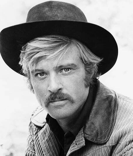 A black and white close-up photo of Mr. Redford in a cowboy hats and corduroy coat. Mr. Redford has a mustache and unruly hair emerging from under his hat.