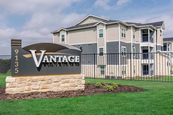 Signage for Vantage at Far Oaks, a residential apartment community, featuring the name prominently displayed. The sign is situated in front of a well-maintained green lawn and a multi-story building with beige siding. There is a fence surrounding the property, and the sky is partly cloudy.