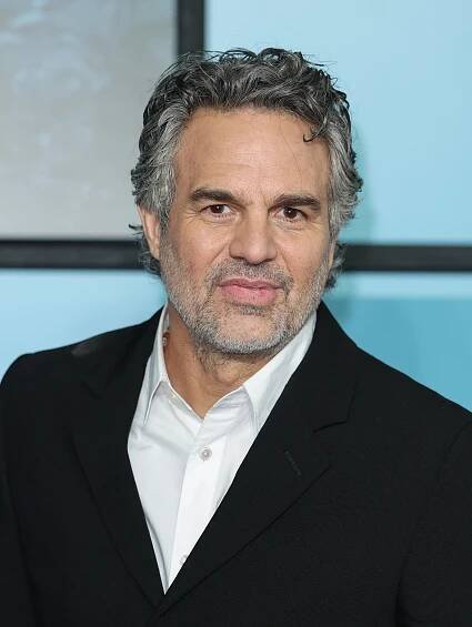 Mark Ruffalo in a black suit and white shirt stands indoors, looking at the camera with a neutral expression
