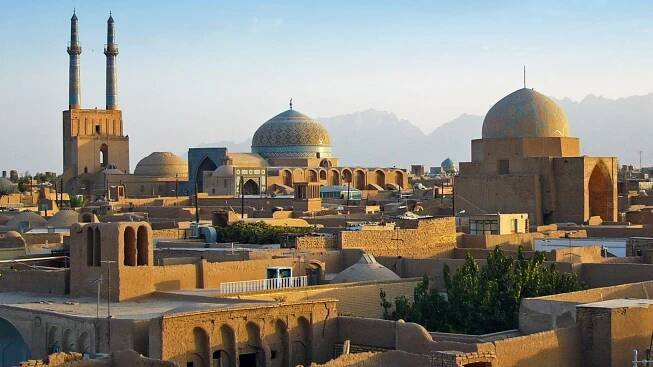 Blick auf die Stadt Yazd im Iran mit traditionellen Moscheen und Dächern, umgeben von Bergen.