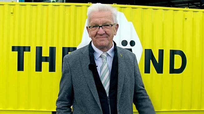 Ministerpräsident Winfried Kretschmann steht vor einem neongelben Container mit "THE LÄND" Schriftzug.