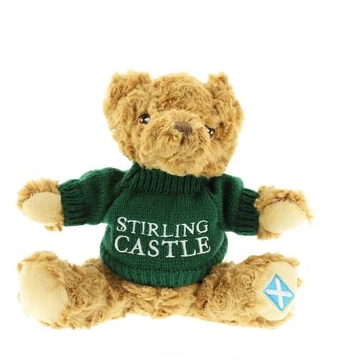 Small cuddly teddy bear wearing a green jumper with white text that reads Stirling Castle. The bear has a St Andrew's flag on it's left paw.  - Image