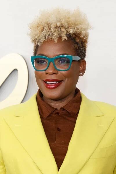 Person with short curly hair, wearing teal glasses, a bright yellow jacket, and brown shirt, smiles at a celebrity event