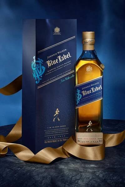 Vedle modré krabičky stojí láhev whisky Johnnie Walker Blue Label, obě zdobí zlaté akcenty. Kolem základny je na tmavém, texturovaném povrchu s modrým pozadím omotaná zlatá stuha.
