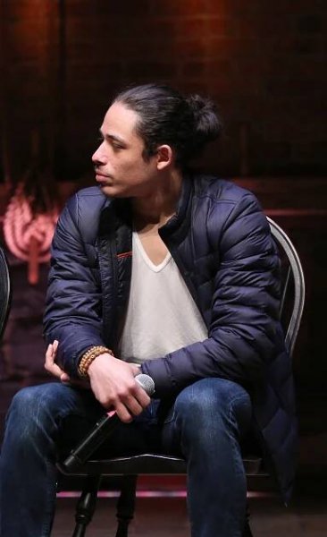 Anthony Ramos seated on stage, wearing a dark jacket and jeans, holding a microphone