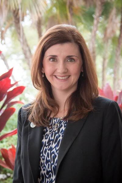 A professional portrait of a woman smiling, wearing a black blazer over a blue patterned blouse, standing outdoors with tropical foliage background.