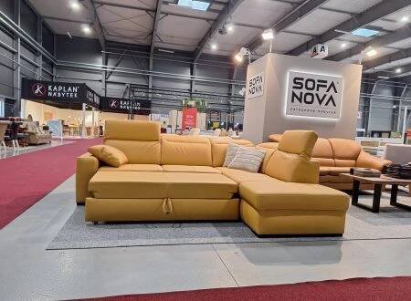 V prostorném showroomu je vystavena velká žlutá sekční pohovka s všívanými polštáři, položená na koberci. Vedle jsou další pohovky a nábytek. V pozadí jsou vidět nápisy "SOFA NOVA" a "KAPLAN".