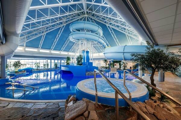 Indoor water park with a winding blue slide above a large pool. Surrounding the area are plants and stone pathways under a high, triangular glass ceiling.