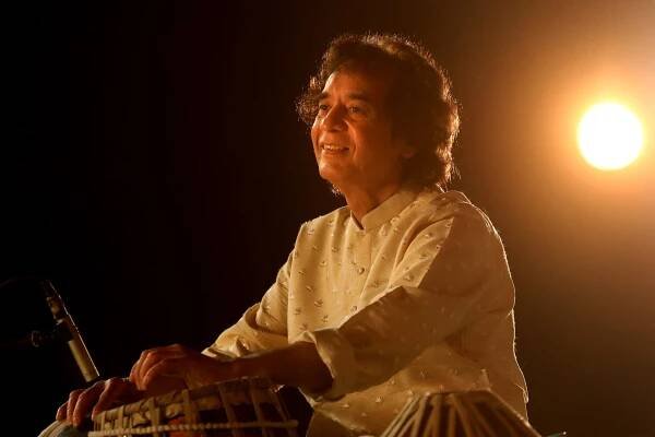 Zakir Hussain wearing a traditional Indian shirt and playing the tabla drum. The sun is in the background. 