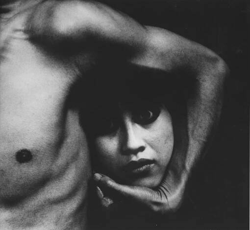 A black-and-white photograph of the left side of a man’s bare torso, his arm around what looks like the disembodied head of a woman.